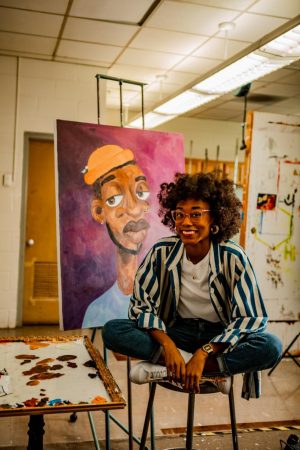 Artist Ebony Johnson Wright sits in front of her painting, "Portrait of Jamal." She primarily paints Black figures to showcase the beauty and joy of Black individuals rather than the pain and discrimination frequently portrayed in art. "I just want them to look like they are important," Wright said about her portraits. "Like they mean something to the world." Wright's work has been featured in many galleries, including at UCM, and she has also won multiple awards for her art and the messages it covers.