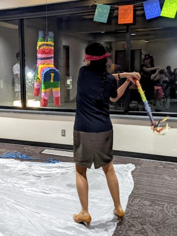 Sandra Merrill is facing away from the camera. She is blindfolded with a bandanna, is wearing a dark short sleeved shirt and shorts. She is swinging a colorful stick and is about to hit pinata.