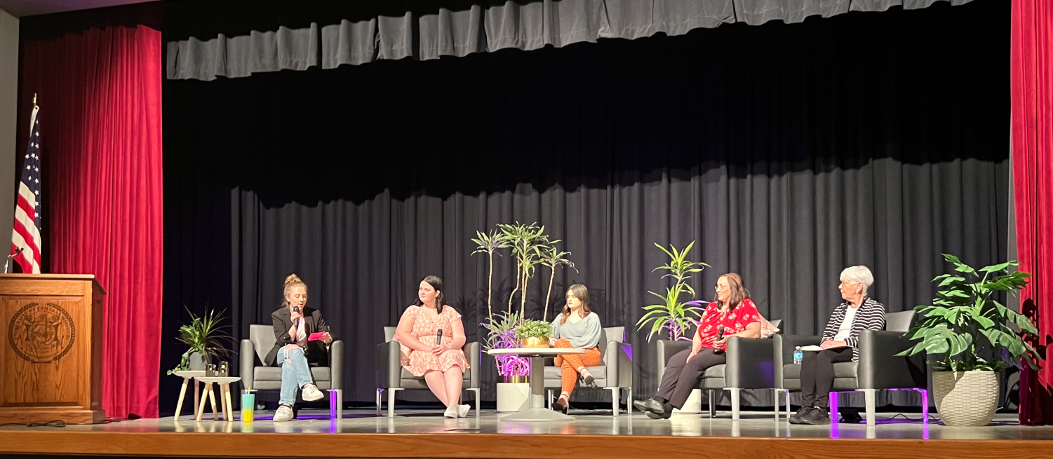 This photo is of a stage with red curtains on the sides and black curtains in the middle. From left to right: there's an american flag behind a wooden podium. There are five women seated on black cushioned chairs and there's assorted plants set up across the stage for decoration. 